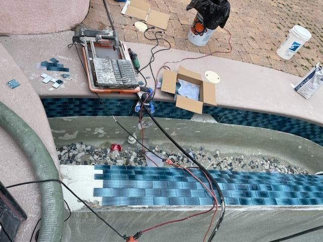 Pool construction: blue tile being installed around the edge, tools and materials scattered nearby.
