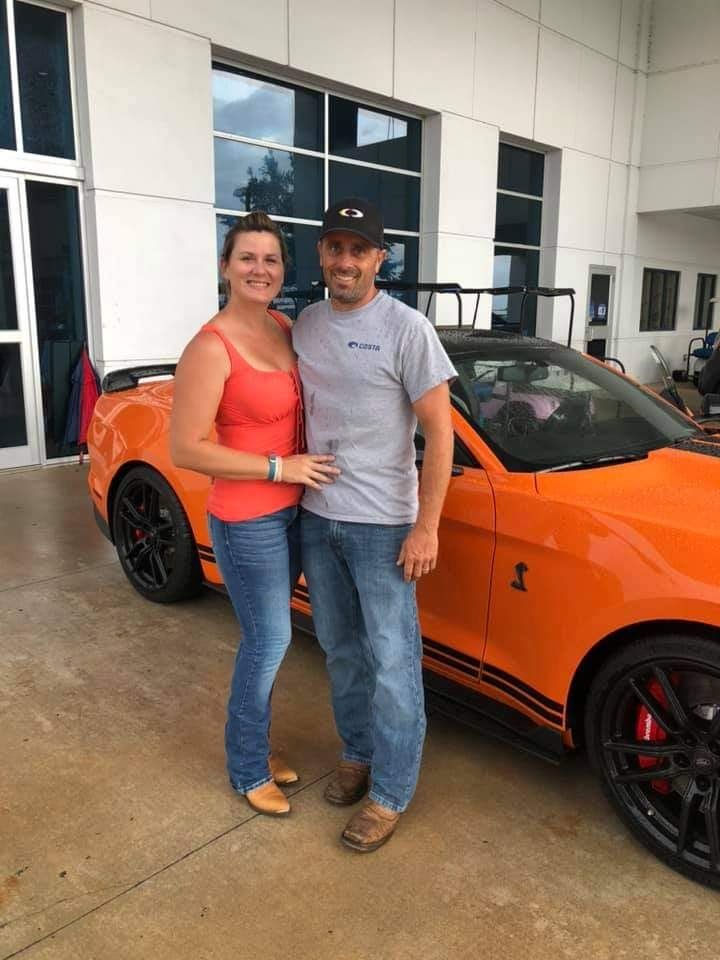 A man and a woman are standing in front of an orange mustang.