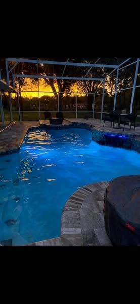 A large swimming pool is lit up at night with a sunset in the background.