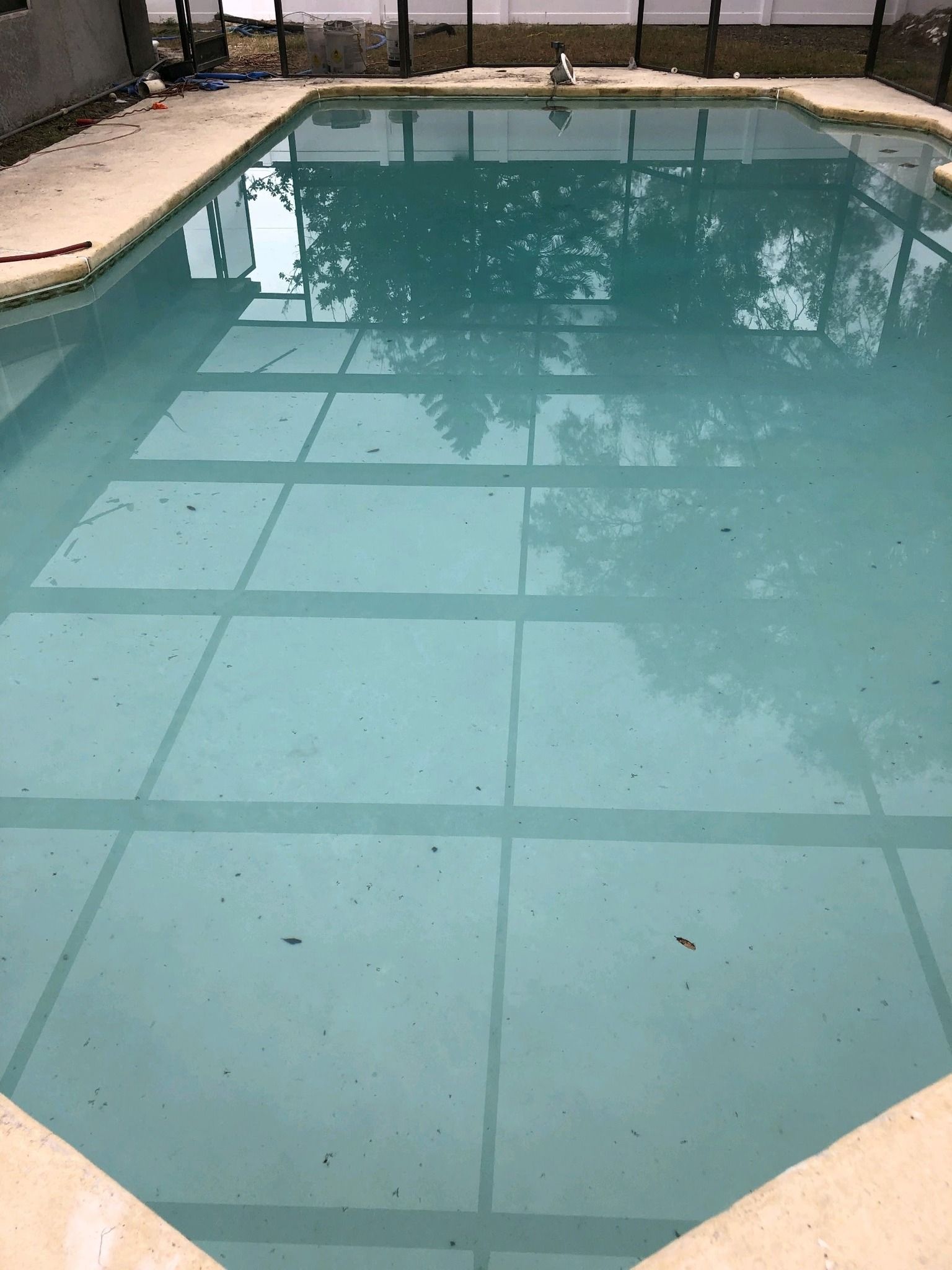 A rectangular pool filled with clear water, surrounded by concrete and a fence; trees are reflected on the water's surface.