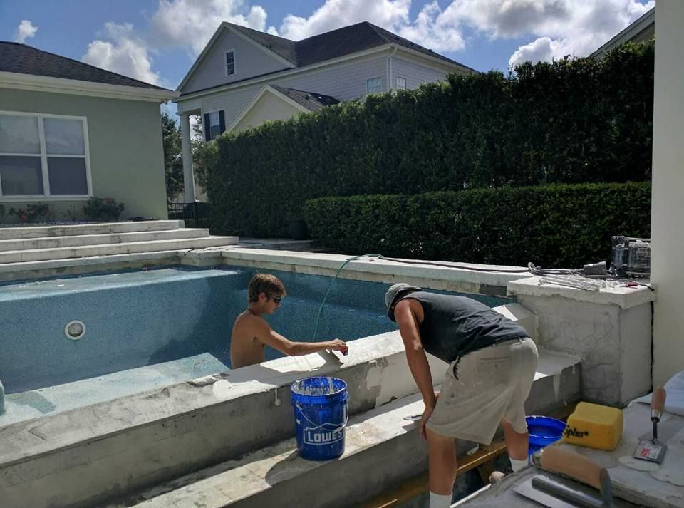 Two people working on a swimming pool: one in the pool, the other outside. Blue pool, white house, and green hedge in the background.