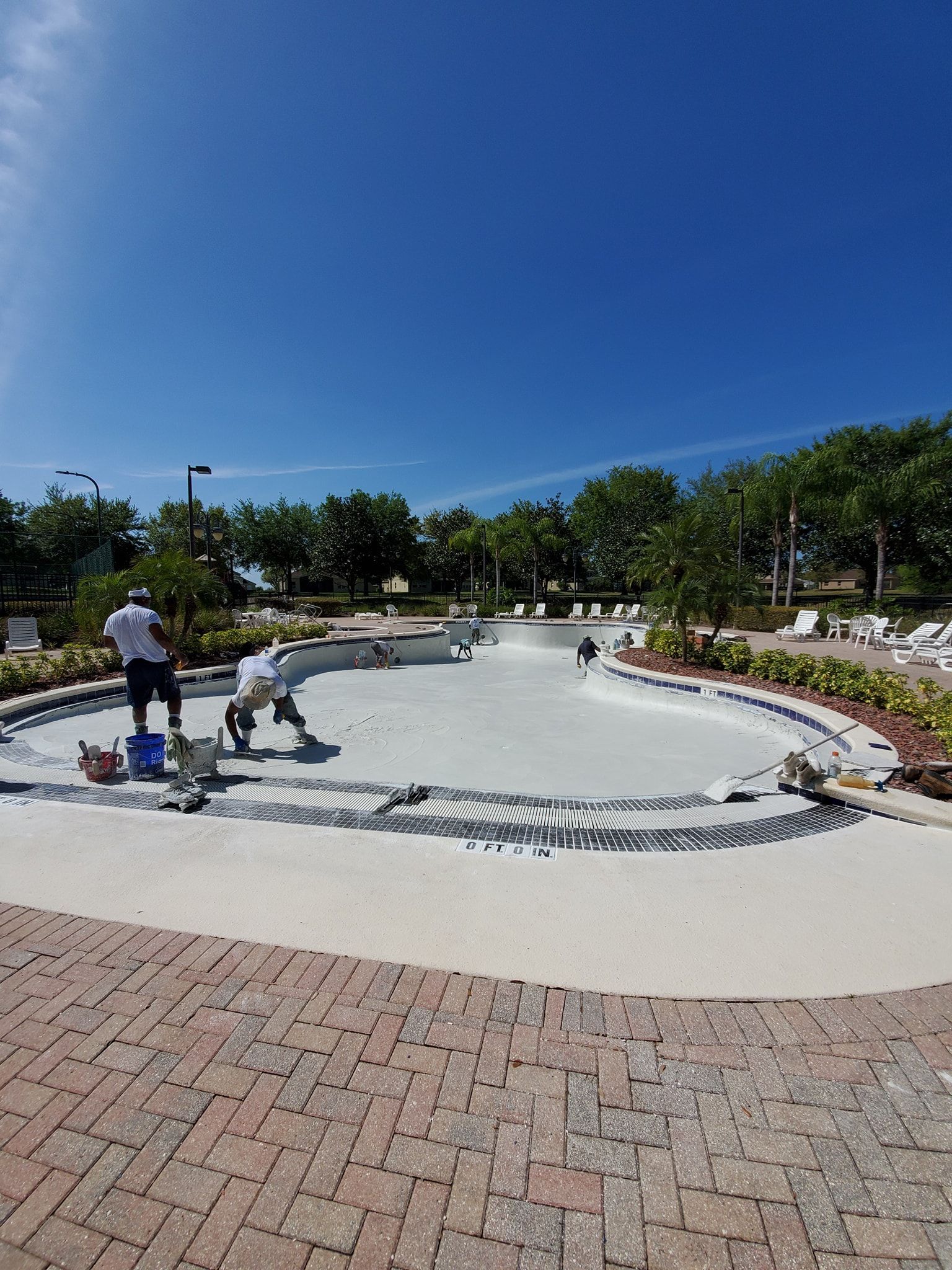 Workers resurfacing a drained pool on a sunny day. Brick patio in foreground, trees and blue sky.