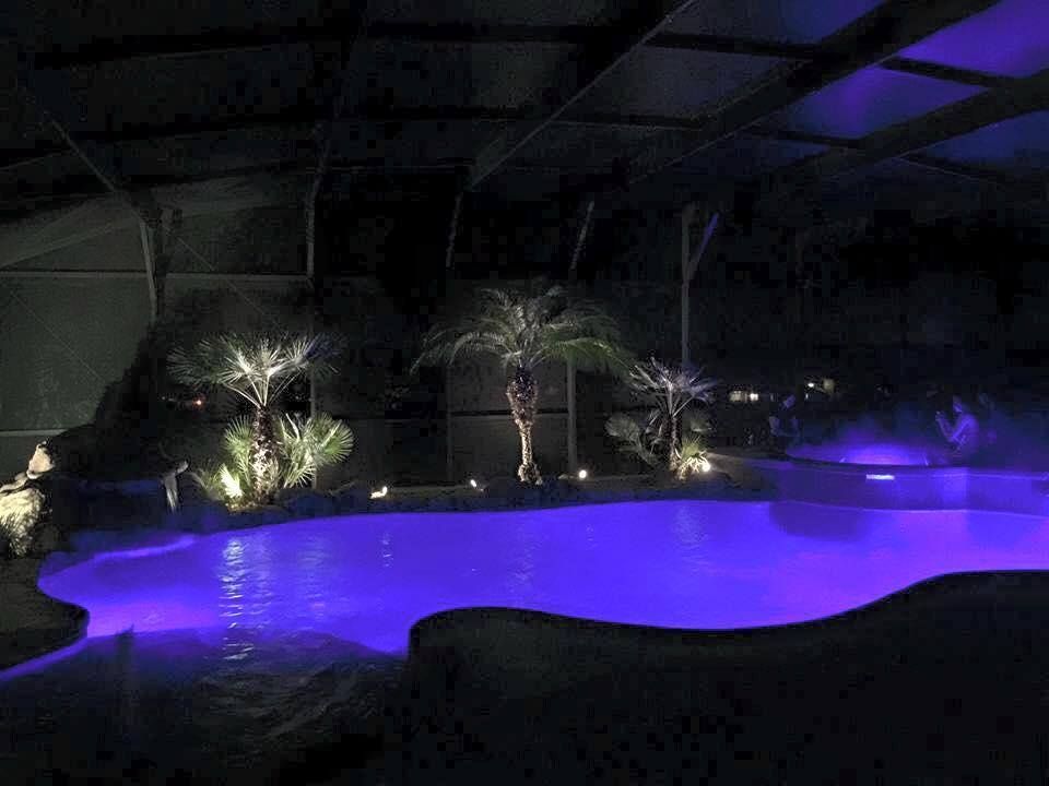 A large swimming pool is lit up at night with purple lights.