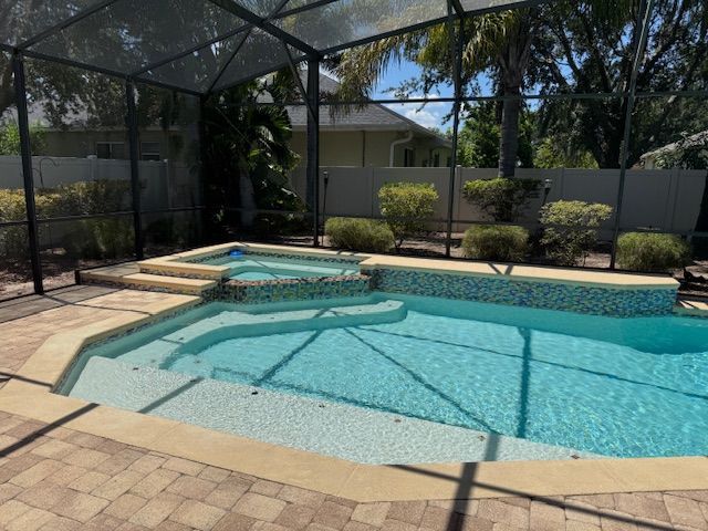 Pool and hot tub within screened enclosure; beige brick patio, blue water, tropical plants.
