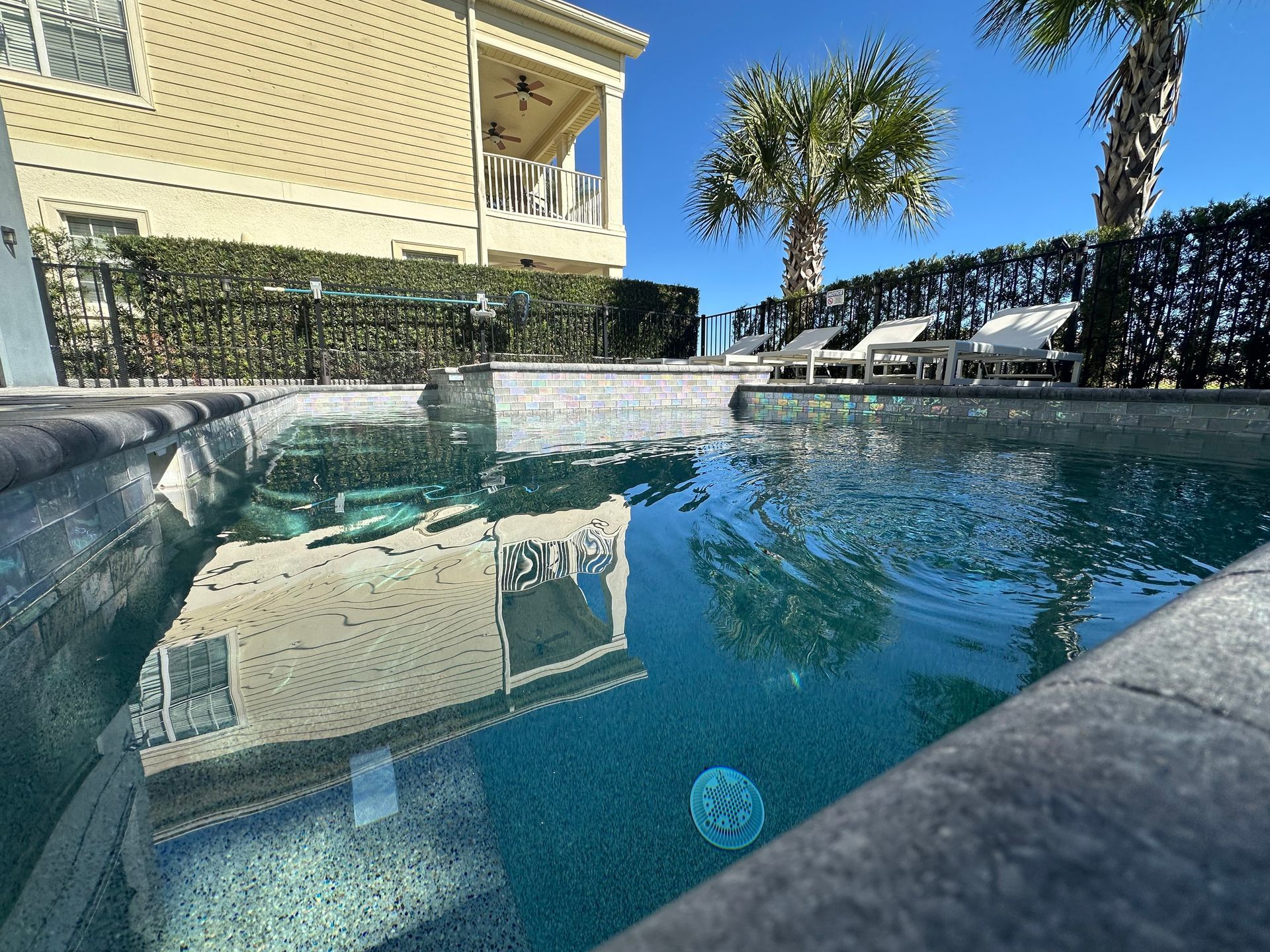 Pool with house and palm trees reflected in the water, under a sunny sky.