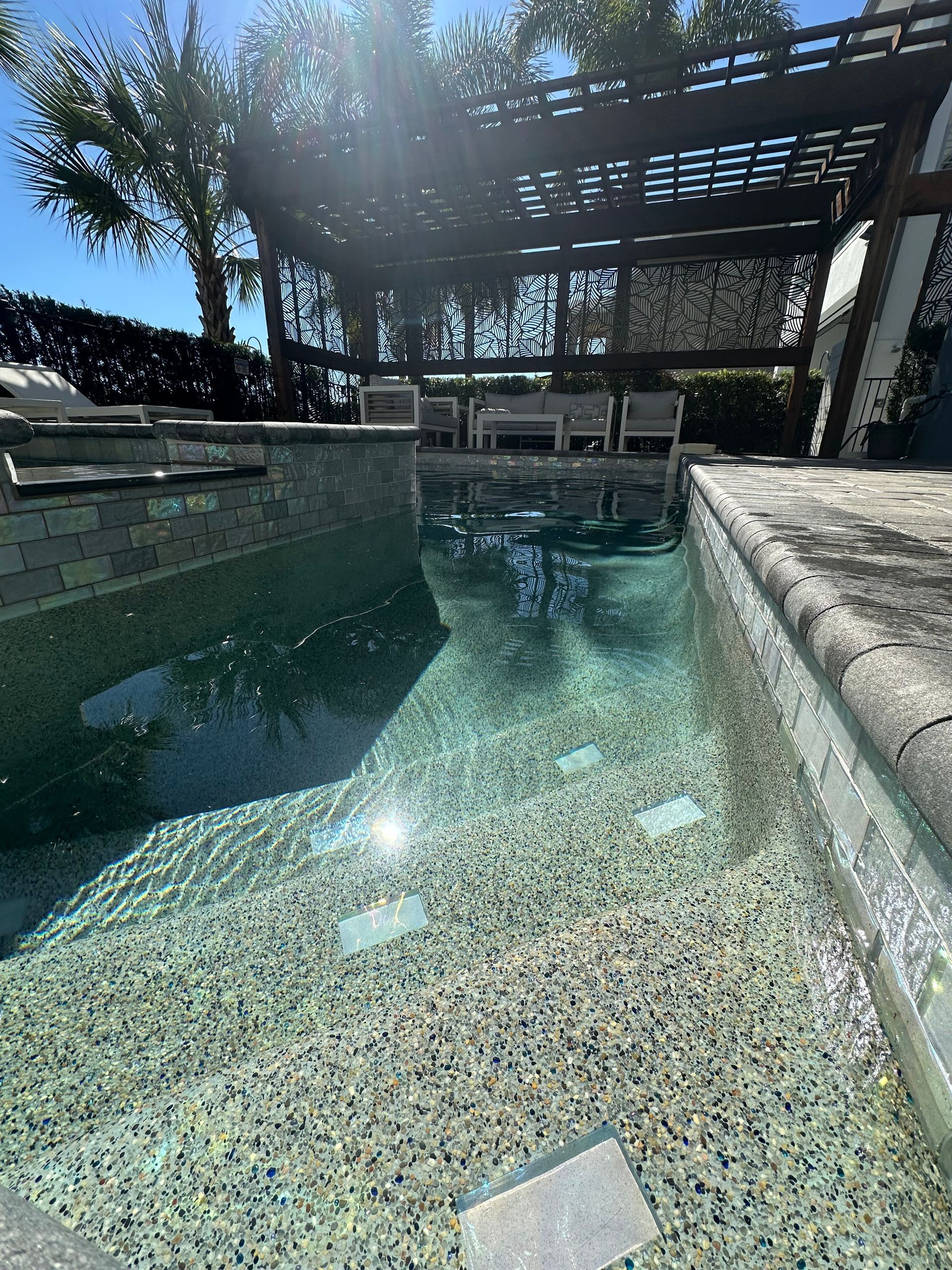 A clear, turquoise pool with steps, facing a seating area under a pergola. Sunlight reflects off the water.