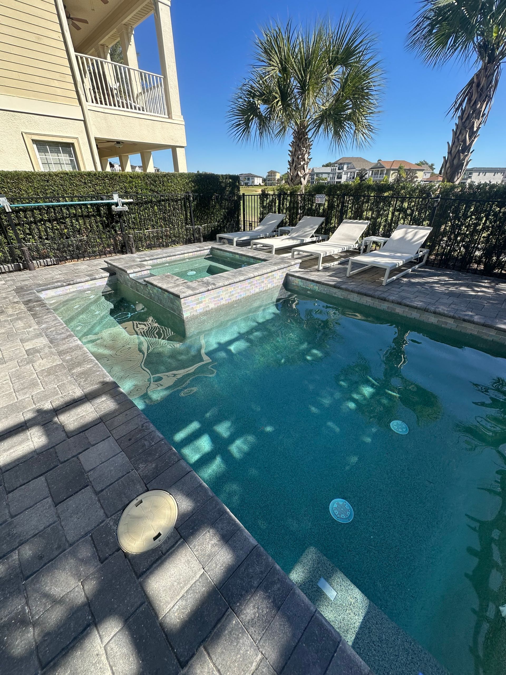 Pool with jacuzzi, surrounded by brick patio, lounge chairs, and hedges, on a sunny day.