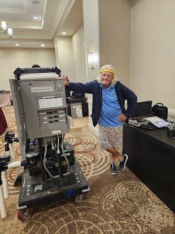 Man in casual clothes leans against a large machine on wheels inside a room with patterned carpet.