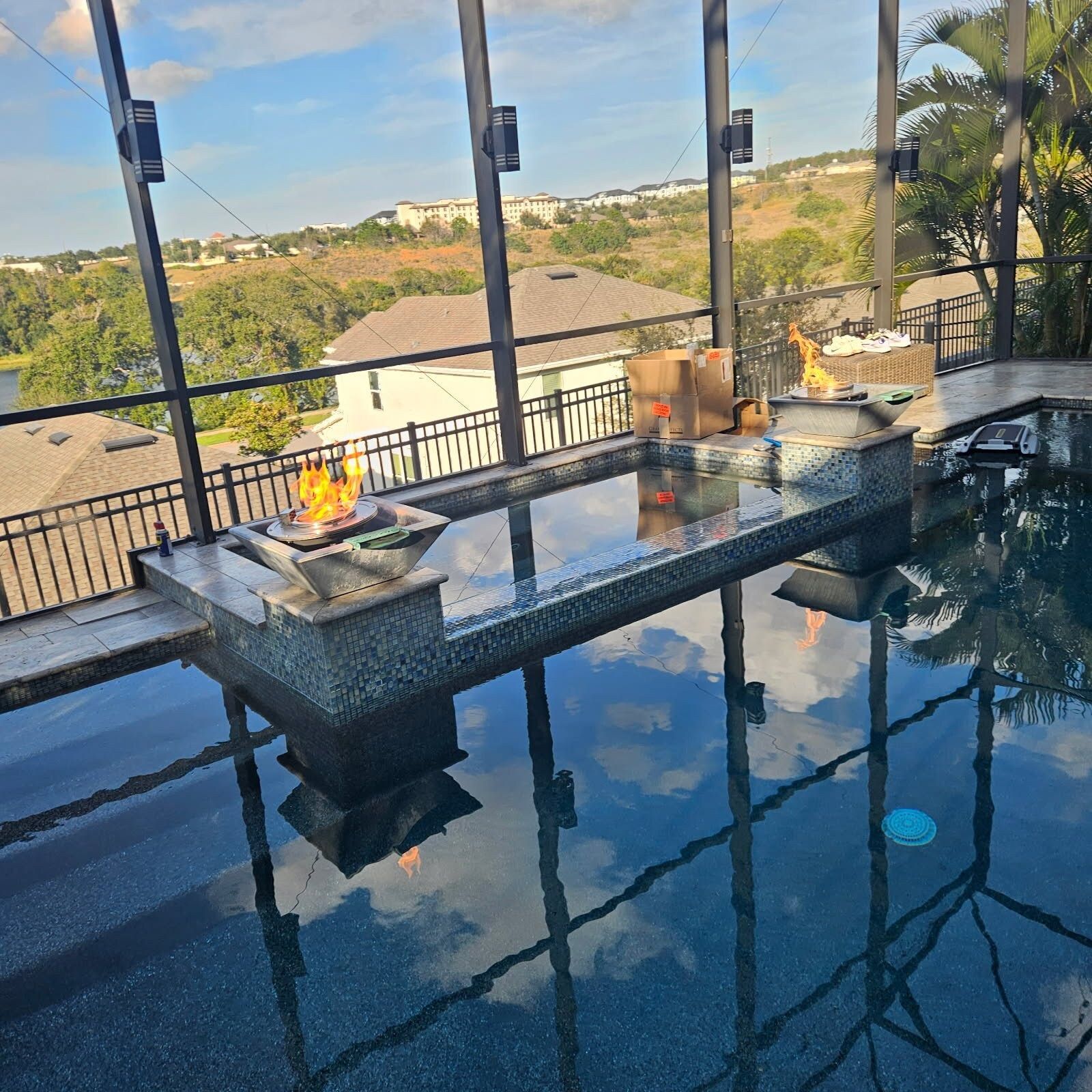 Pool with fire pit, reflecting blue sky, surrounded by black railing and glass panels, overlooking a landscape.