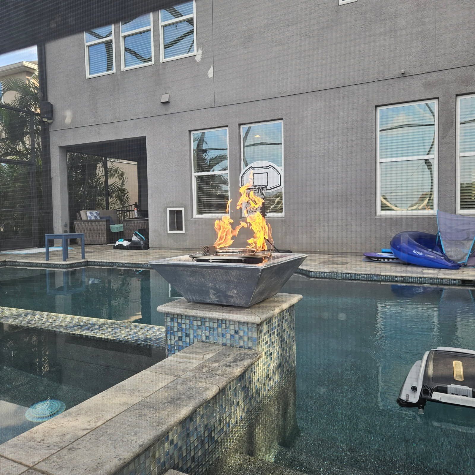Backyard pool with fire pit, basketball hoop, and house. Blue water and gray walls.