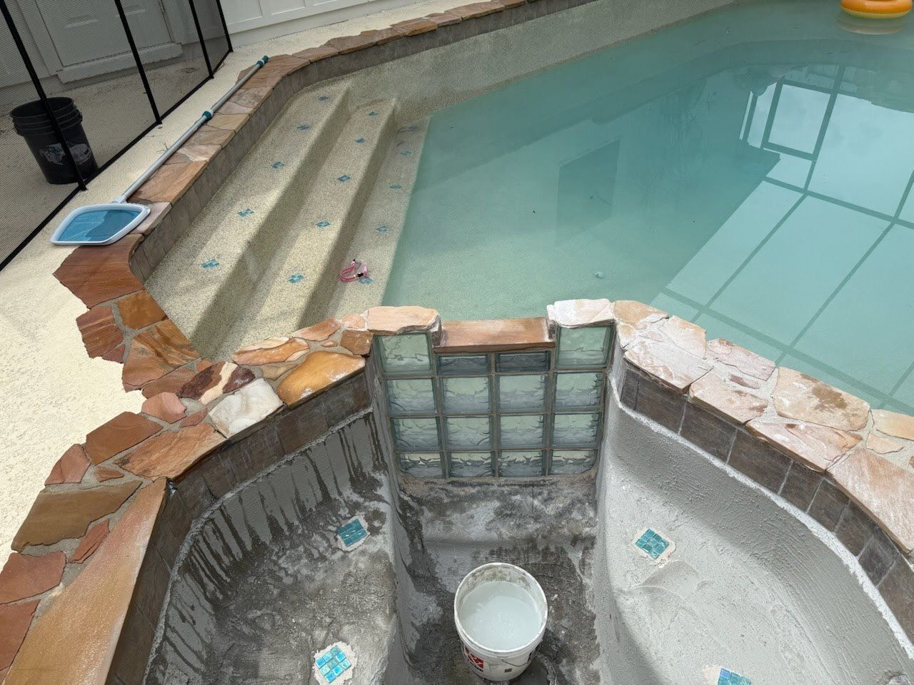 Pool under construction with brick and tile surround; steps, a water bucket, and glass block wall are visible.