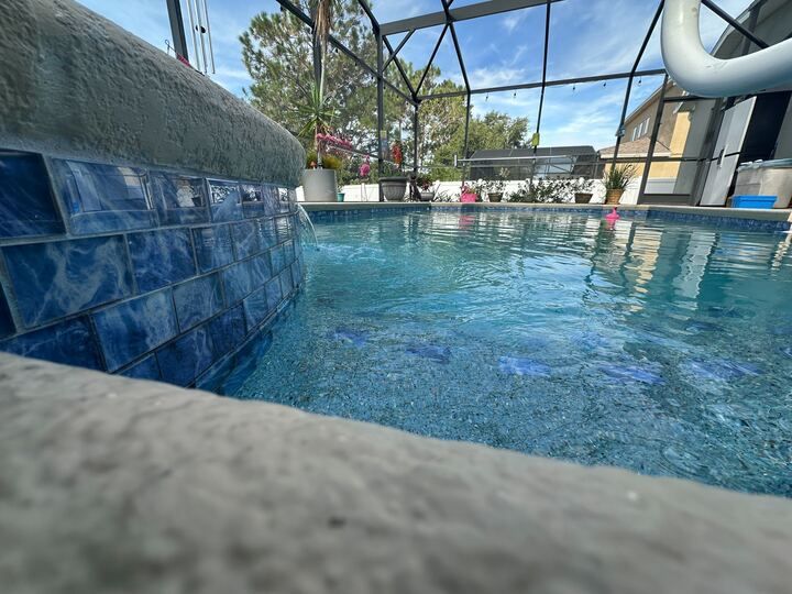 Blue-tiled pool with clear water, bordered by a gray concrete edge, under a screened enclosure.