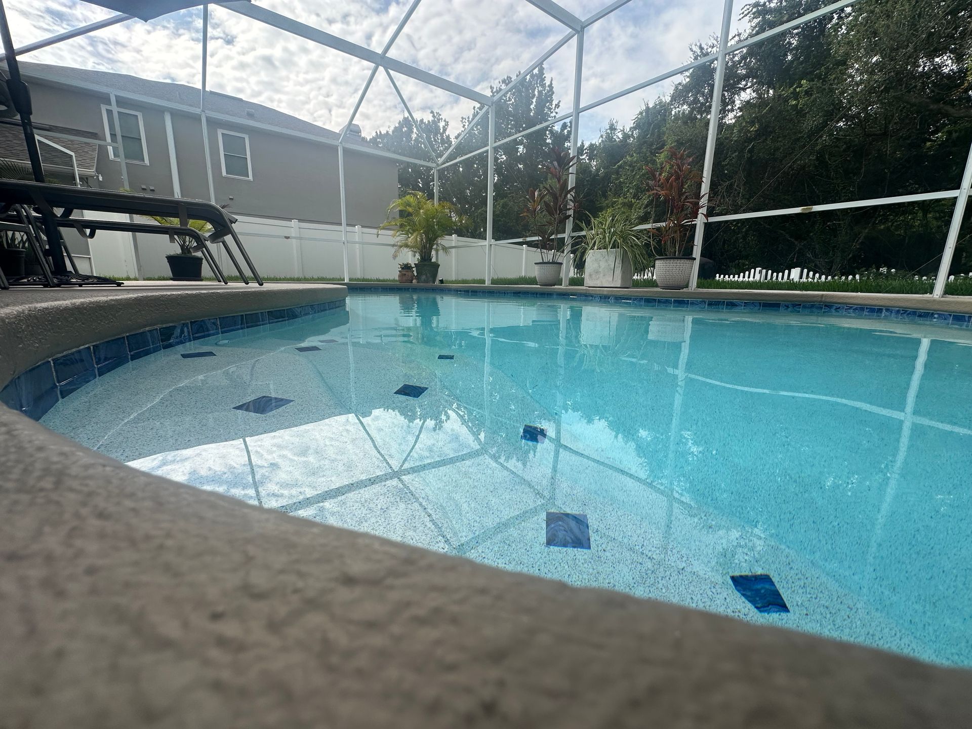 Swimming pool with blue water and tiled edge, enclosed by a screened lanai, next to a house.