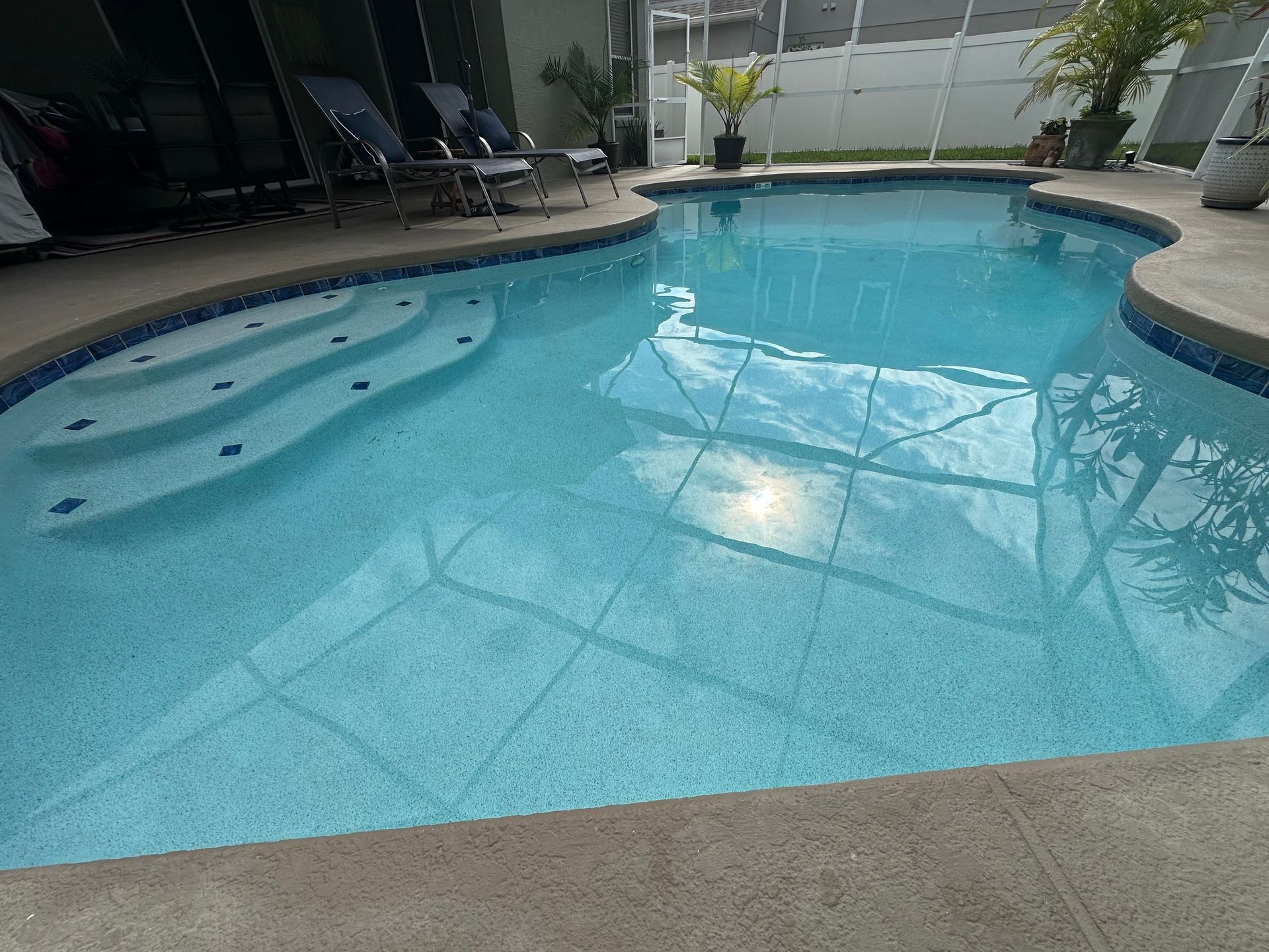 Pool with blue water and steps. Patio chairs sit poolside. Bright sunlight reflected in the water.