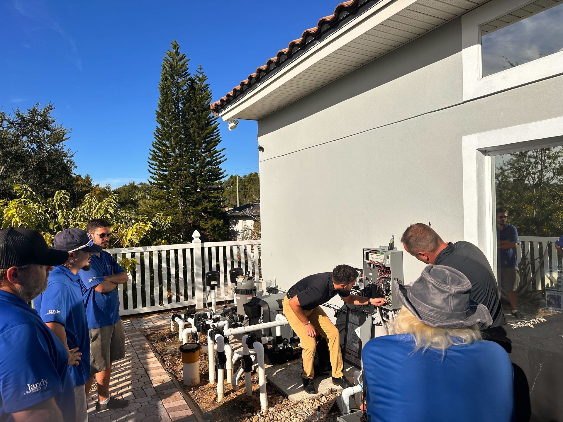 Pool technicians examining pool equipment near a house. Several workers in blue shirts are gathered outside.