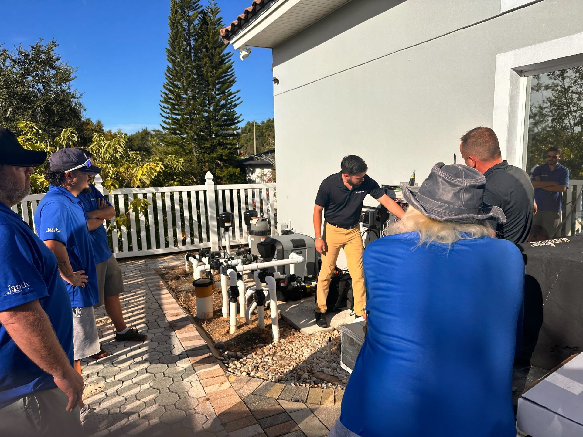 Group of people observing pool equipment outside a house. Some wearing blue shirts. Bright, sunny day.