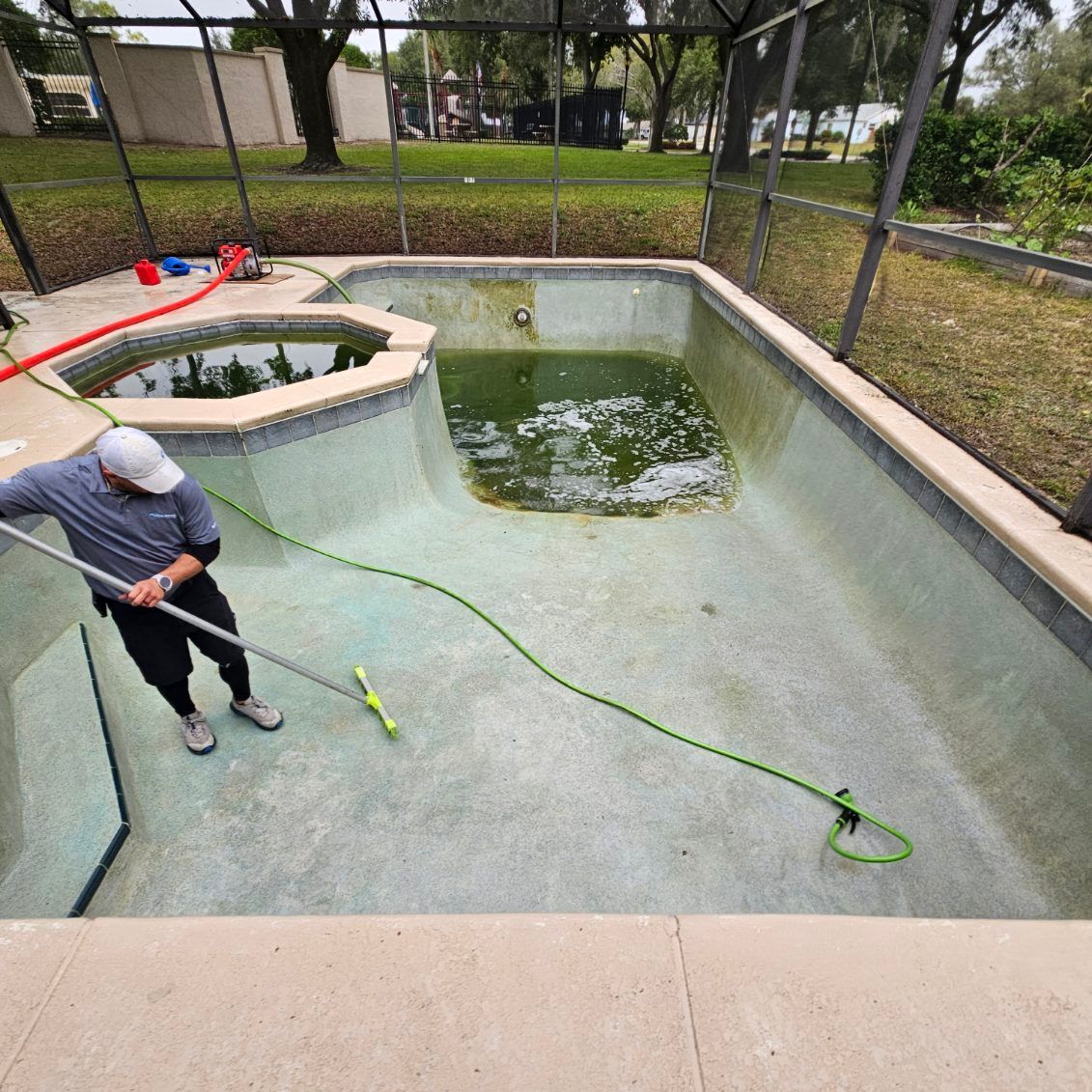 A person cleaning a green, algae-filled pool with a long-handled brush.