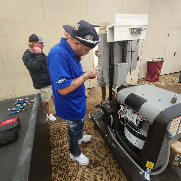 Three people inspecting a machine in a room. One man in a blue shirt examines wiring. Another man stands nearby and looks on.