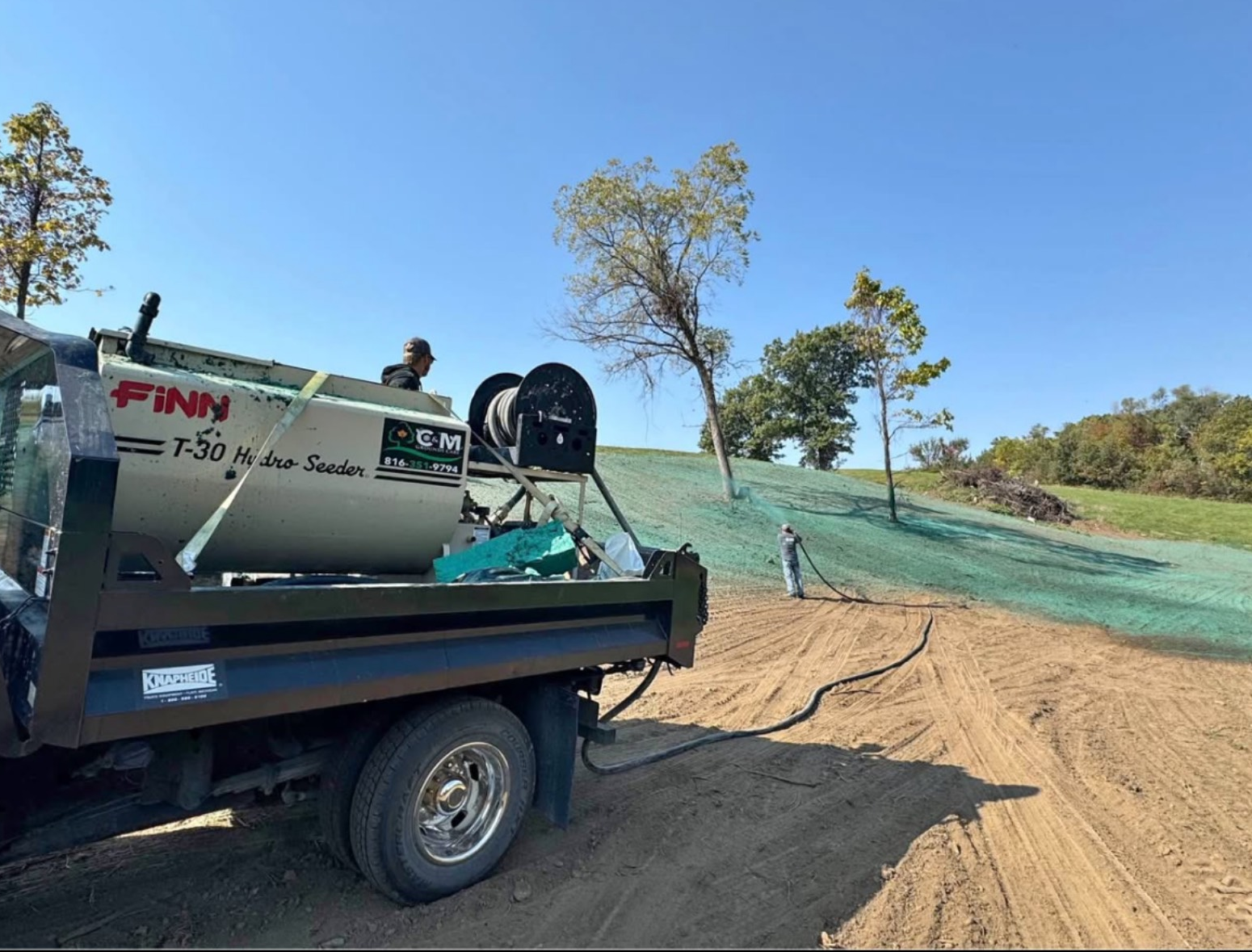 Person spraying green liquid from a hose, possibly for lawn care, on a sunny day.