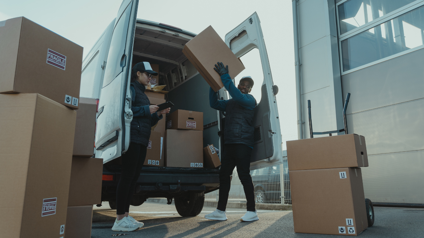 Two movers loading cardboard boxes into a white delivery truck at a warehouse dock