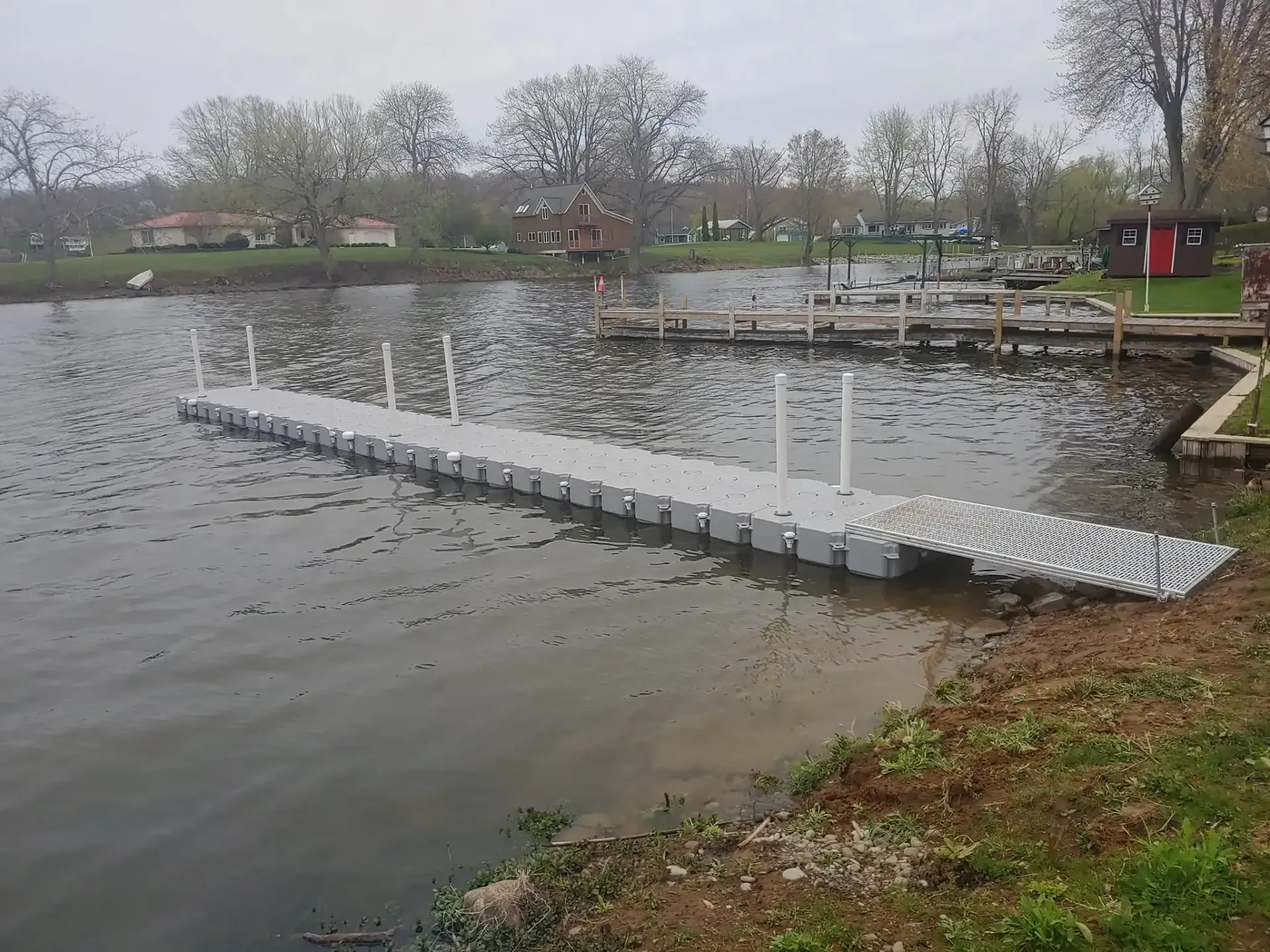 Floating dock in a calm pond, with white posts and a grassy shore under cloudy skies.