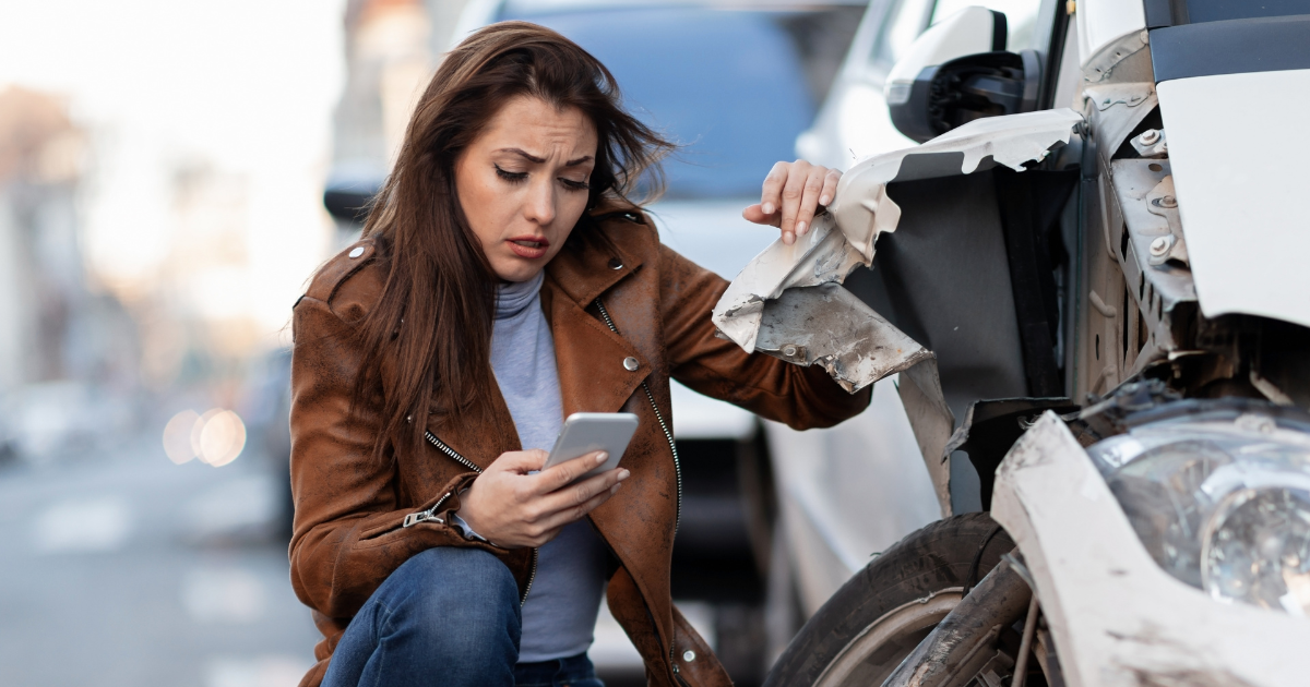 A woman is kneeling down next to a damaged car and looking at her phone.