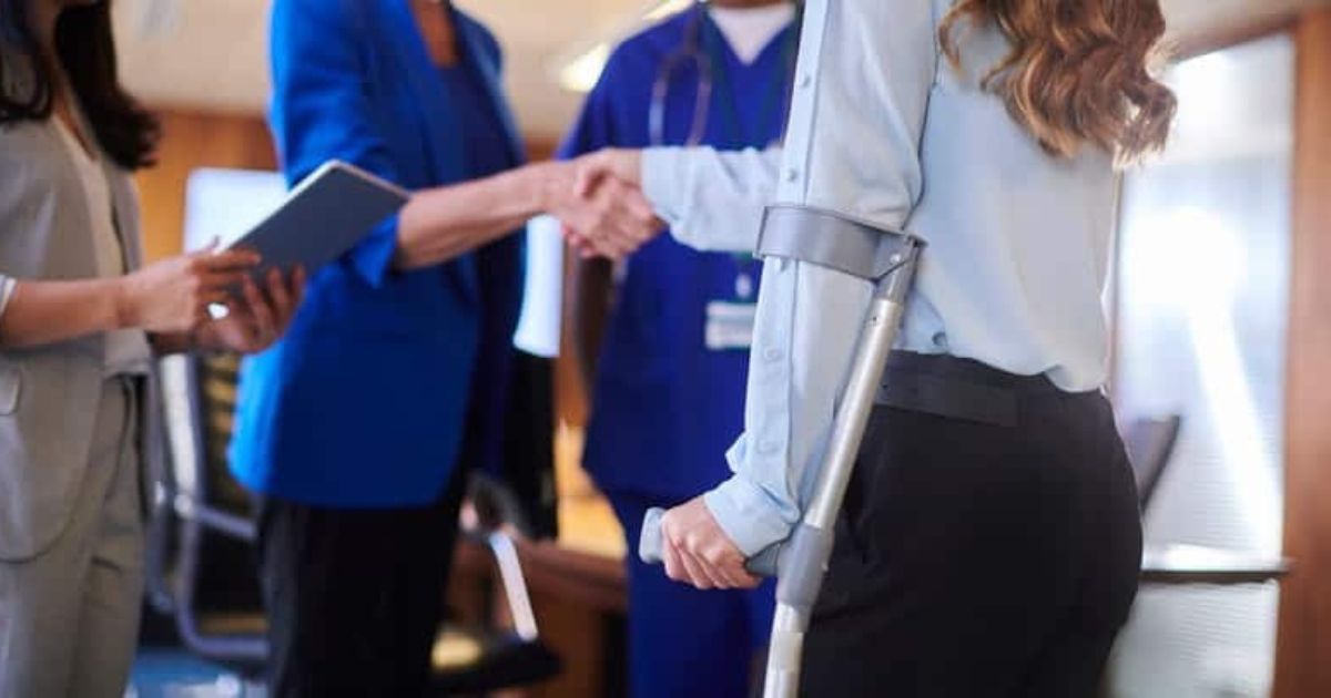 Woman with crutch shakes hands in office setting; other people present.