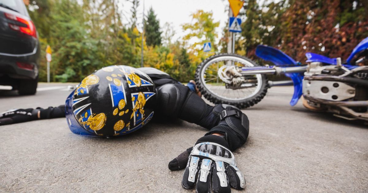 Motorcyclist lying on asphalt road next to a tipped over blue and white motorcycle after accident with a car.