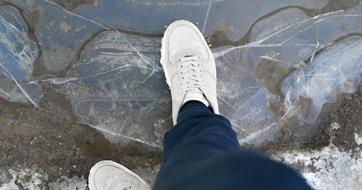Person's foot stepping on cracked ice, wearing white sneakers and dark pants.
