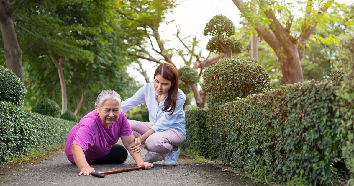 Woman helping elderly person who has fallen on a path, with green bushes and trees in the background.
