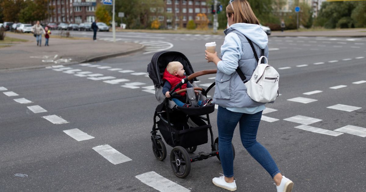 Woman pushing a stroller with a baby across a crosswalk, holding a coffee cup.