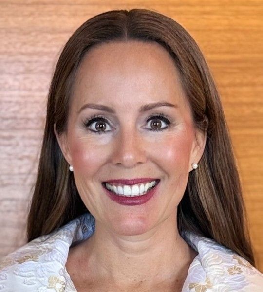 A smiling person with long brown hair, wearing a white textured jacket and small pearl earrings against a wood background.