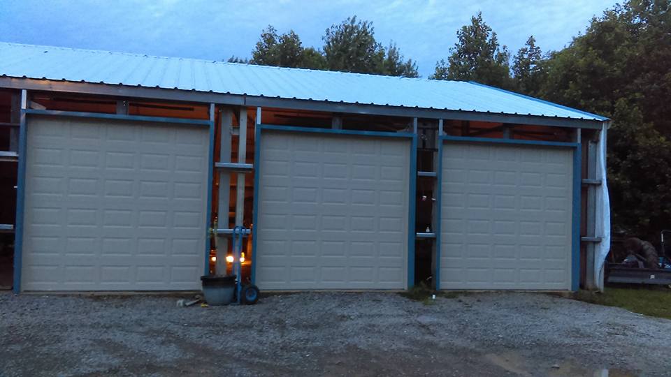 Three-bay garage with light beige doors, blue trim, and metal roof at dusk.