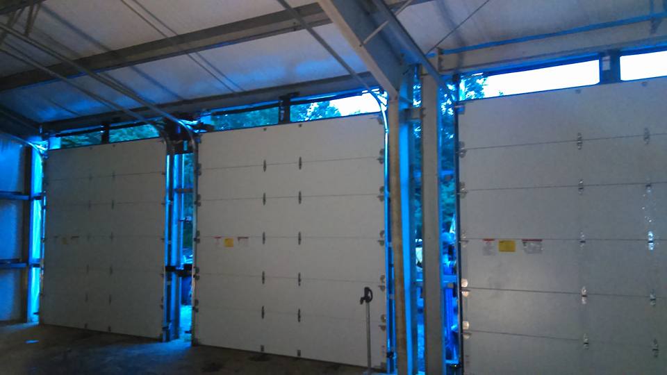 Three white garage doors inside a building, windows above. Blueish lighting.