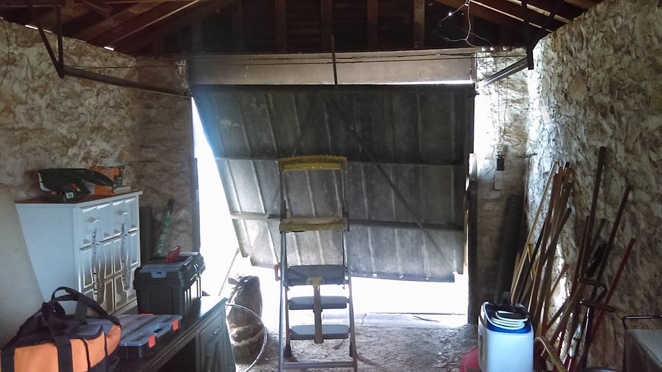 Interior of an old garage with open door, tools, and a stepladder; walls are made of plywood.