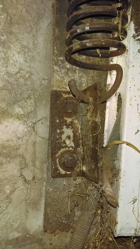 Rusty metal hardware bolted to a concrete wall, with a large coil spring attached.