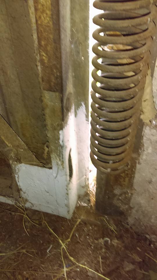 Corner of a weathered wooden structure with a rusty spring visible. White paint accents the corner.