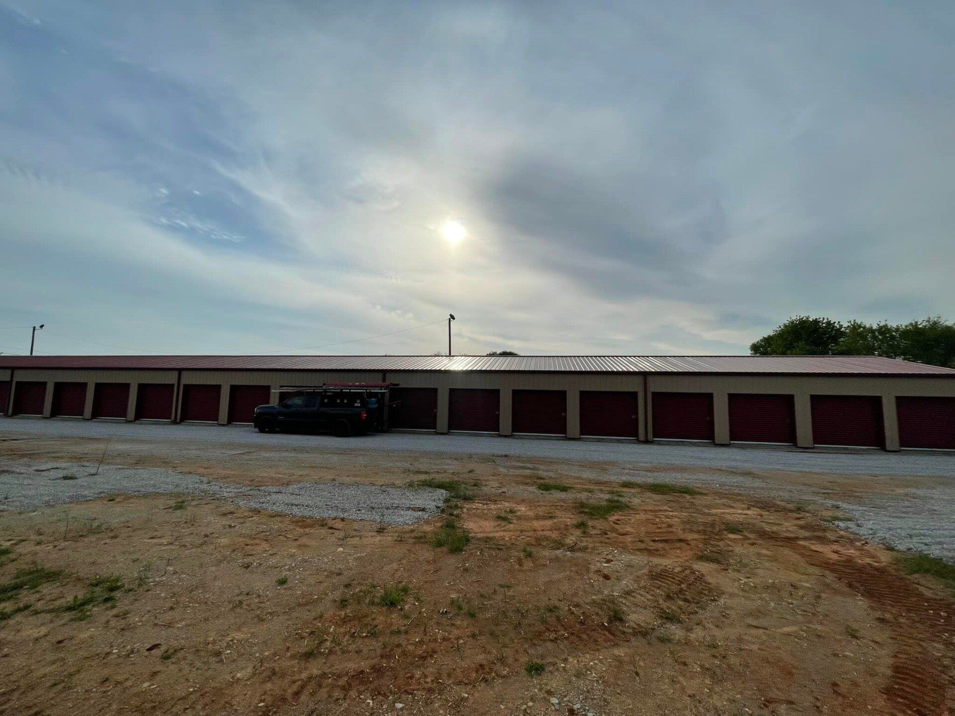 Storage units with red doors and tan roof, with a black vehicle in front, under a bright sun.