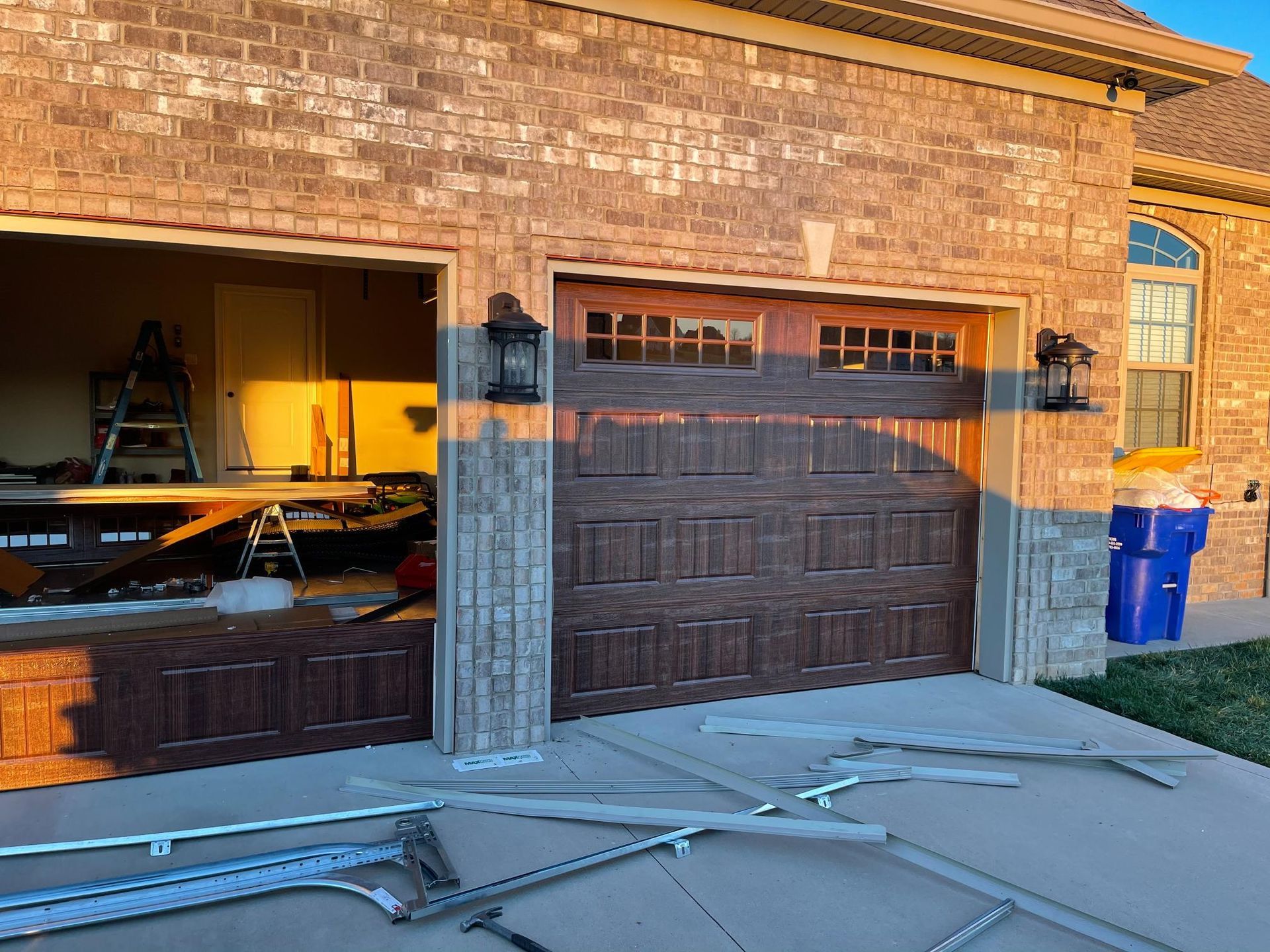 Garage with a brown door and a partially open bay with construction materials. Brick exterior and blue trash can.