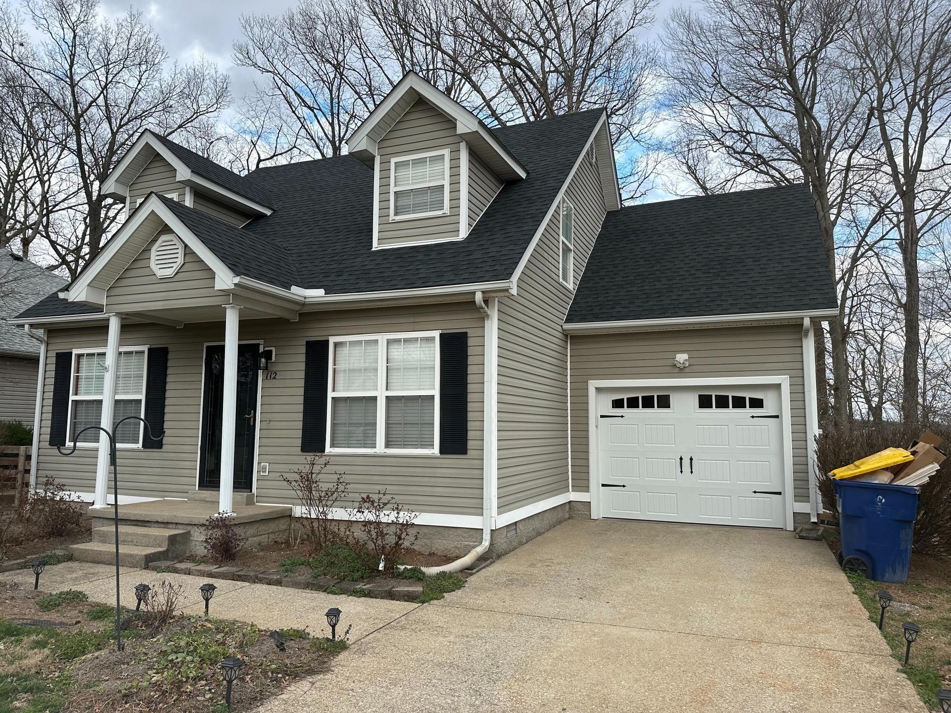 Tan house with black roof, white trim, and a one-car garage. A driveway leads to the garage.