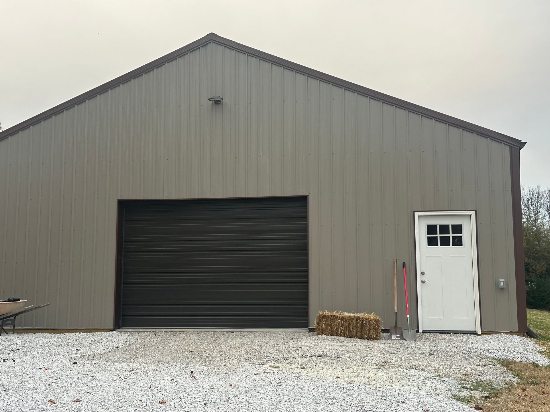 Gray metal barn with a brown garage door and a white door, set on gravel.