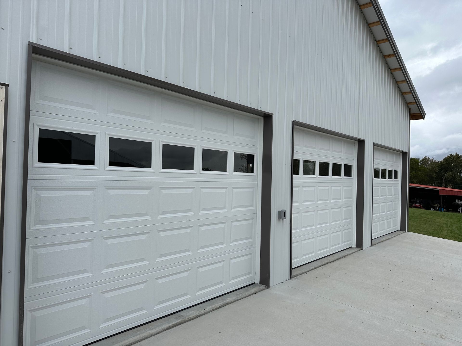 Three white garage doors on a light gray metal building, set on a concrete pad, against an overcast sky.