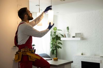 Electrician in red overalls installing a light fixture in a white room.