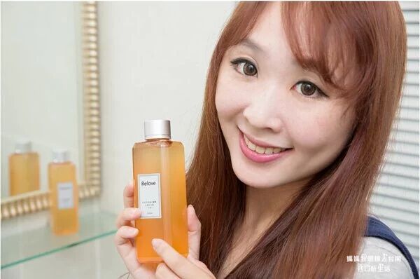 一位女士面帶微笑，手裡拿著一瓶橘色的液體保養品，旁邊是一面鏡子，鏡子裡還有兩瓶類似的保養品。