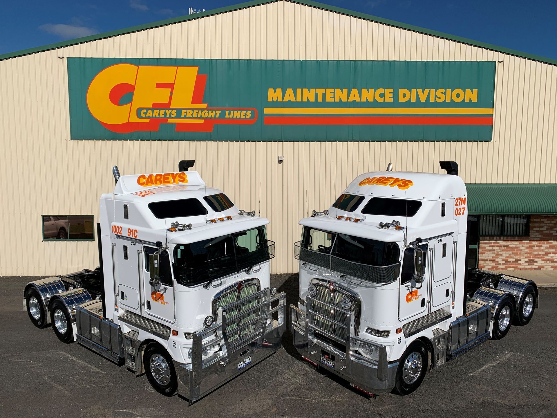Front of a white semi-truck with a metal grill and two headlights, parked next to a trailer in daylight.