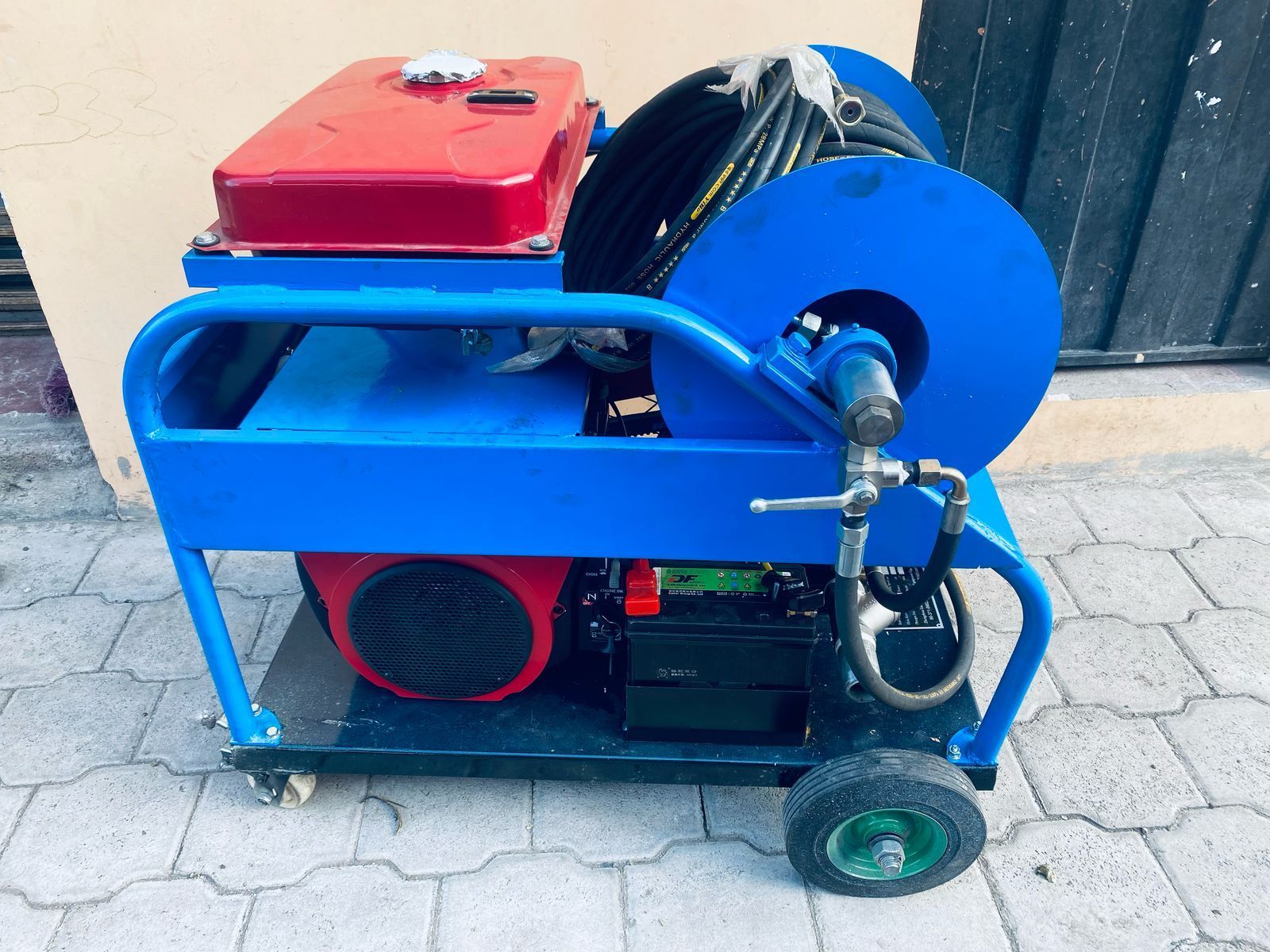 Máquina de metal azul con motor rojo y gran polea azul sobre un bastidor con ruedas al aire libre.
