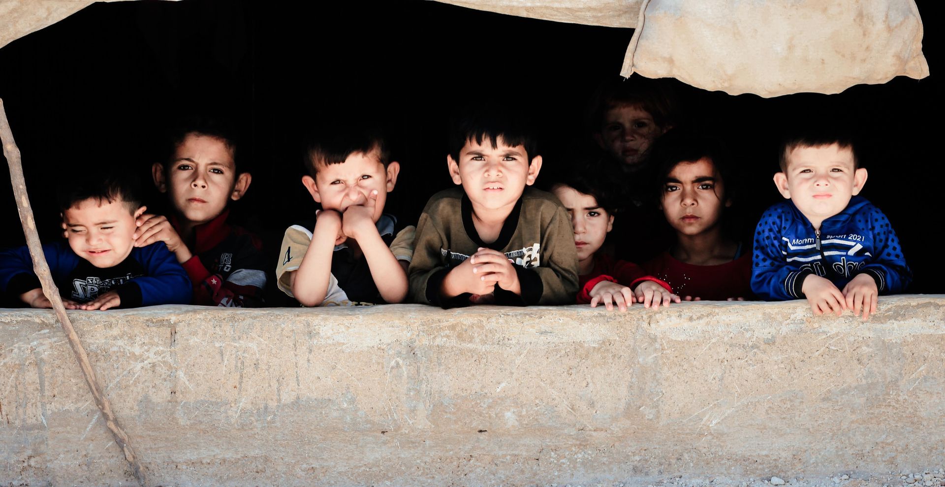 Huit enfants regardent au loin depuis une ouverture sombre en pierre, semblable à une grotte, leurs visages illuminés par une douce lumière.