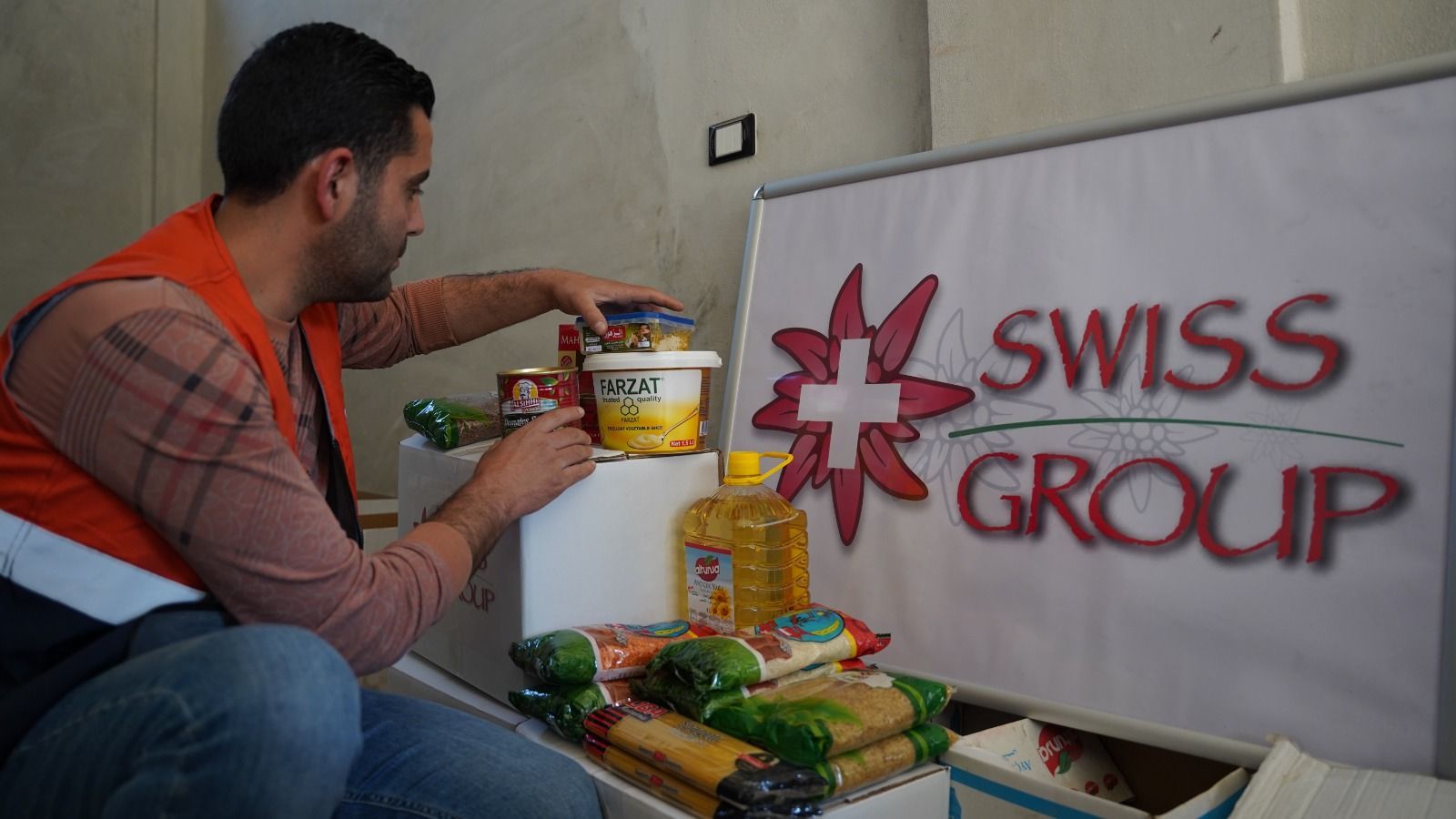 Une personne portant un gilet orange range des aliments dans des boîtes à côté d'un panneau indiquant « Swiss Group ».