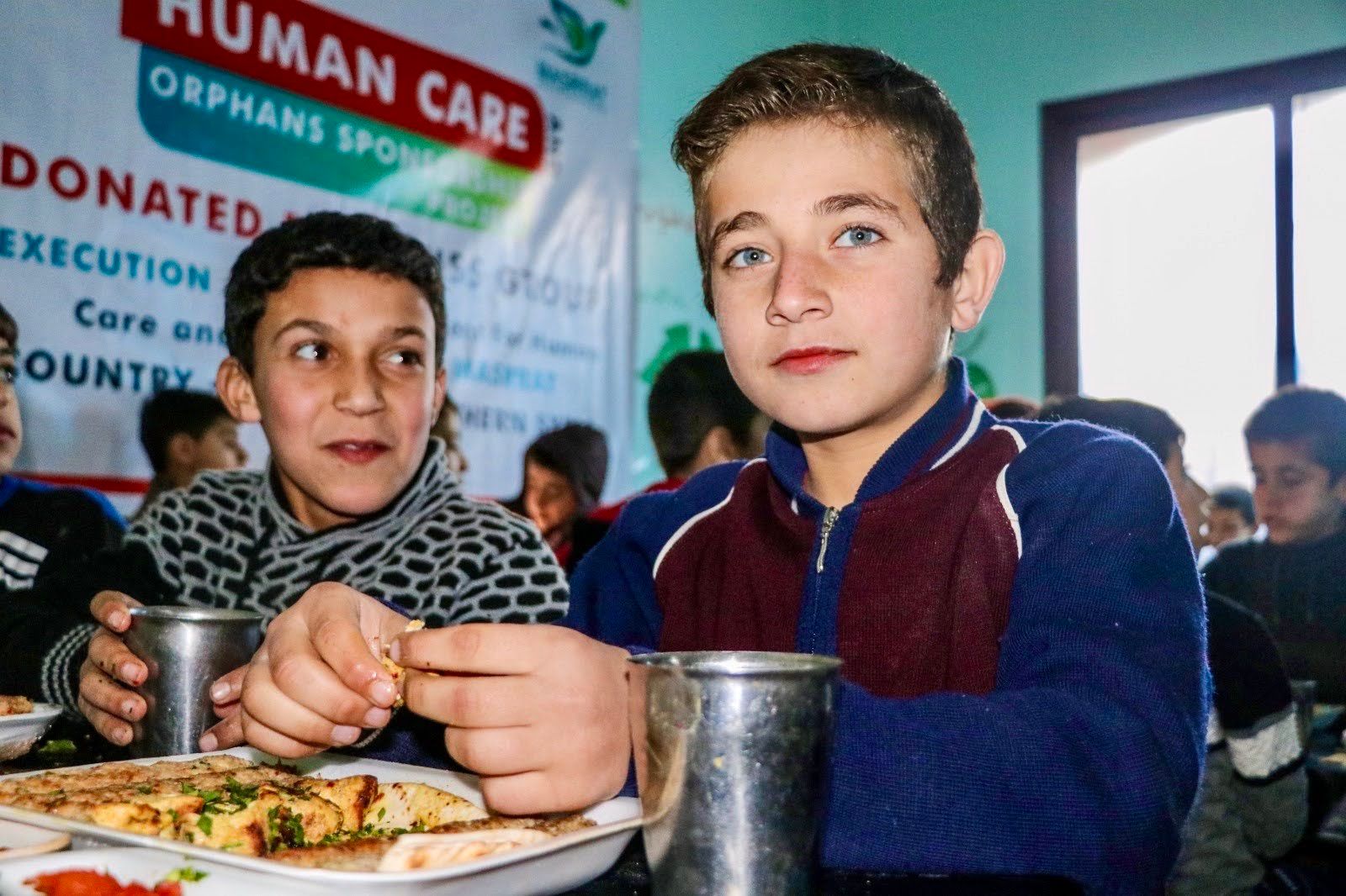 Deux enfants sont assis à une table avec de la nourriture et des gobelets en métal devant une banderole Human Care.