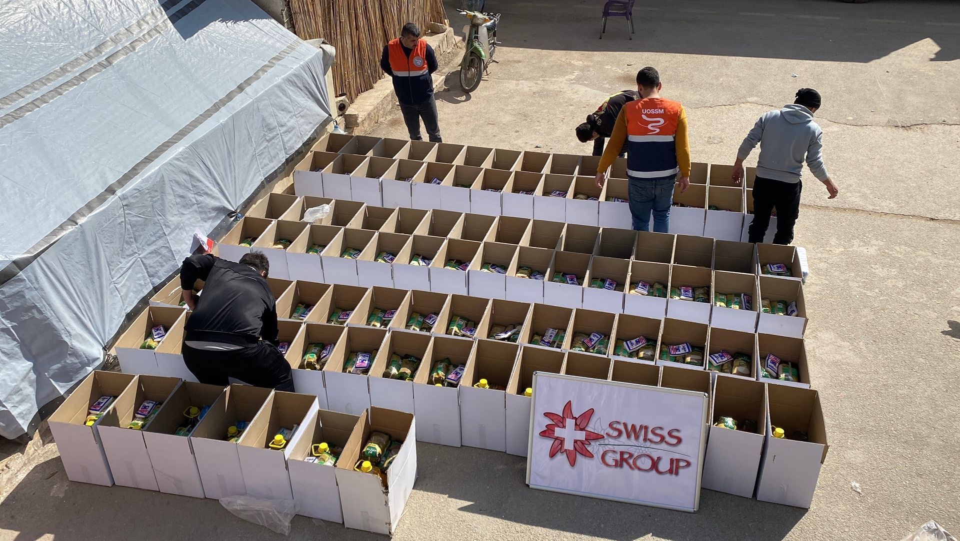 Des bénévoles disposent à l'extérieur des rangées de cartons remplis de fournitures, à côté d'un panneau indiquant « Swiss Group ».